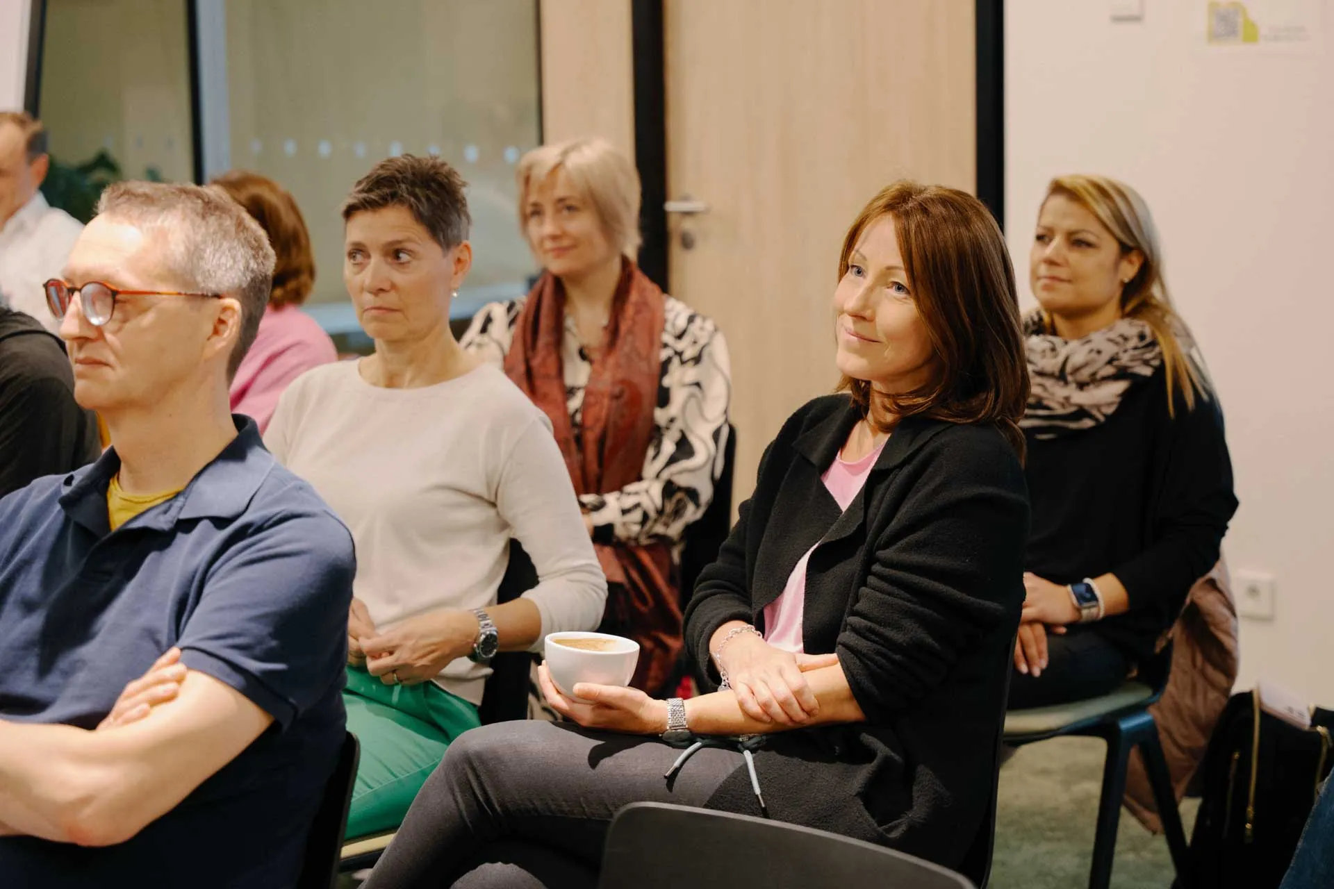 A group of people sitting attentively in a meeting room, with one person holding a cup. They appear engaged and focused on a presentation or discussion.
